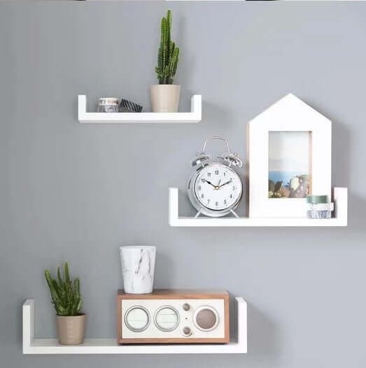 White wood wall-mounted shelf set showcasing decorative items like plants, clock, and photo frame on a gray wall.