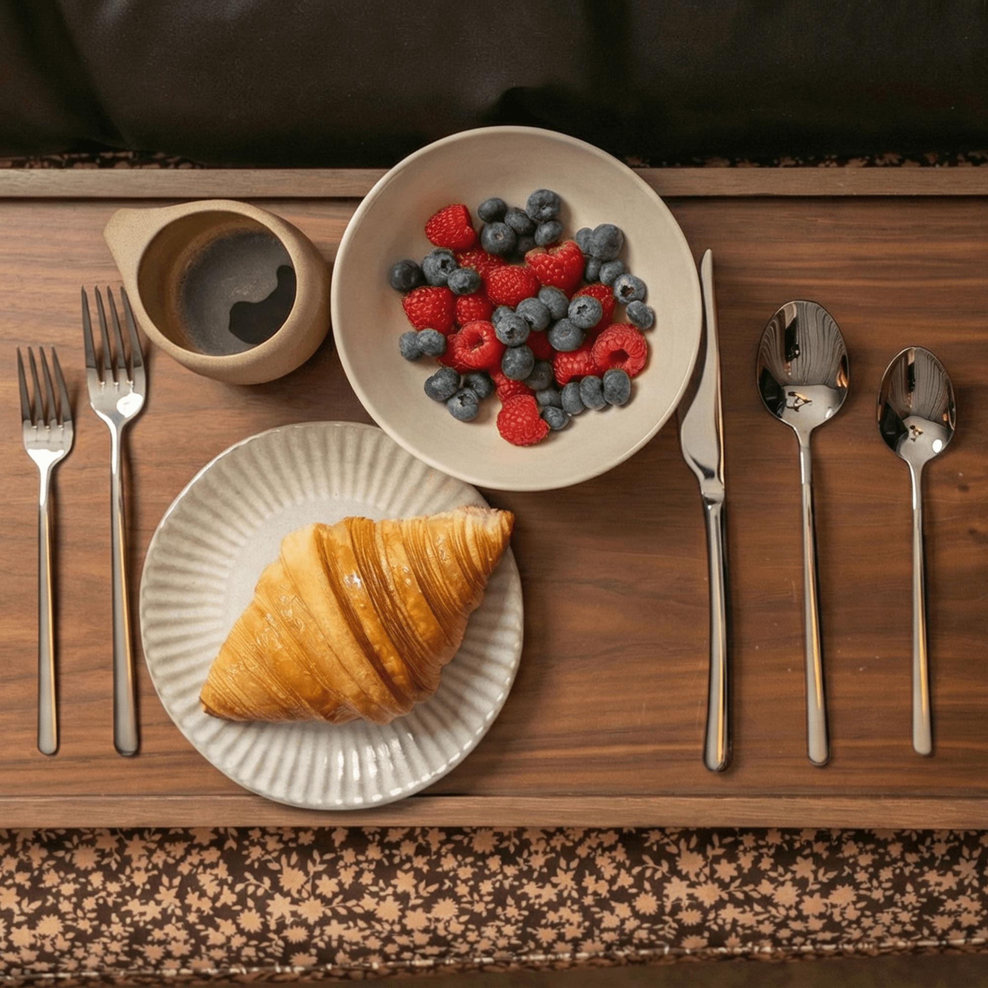 Emilia Mirror 20-Piece Flatware Set displayed with croissant and berries on a wooden table.