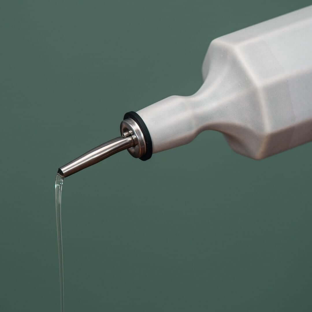 Ceramic olive oil dispenser pouring oil, with a sleek design and stainless steel spout against a muted background.