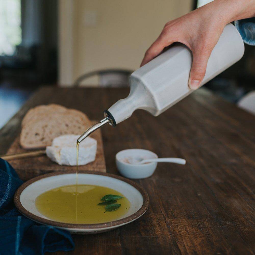 A hand pouring olive oil from a ceramic olive oil dispenser into a plate, with bread and cheese in the background.
